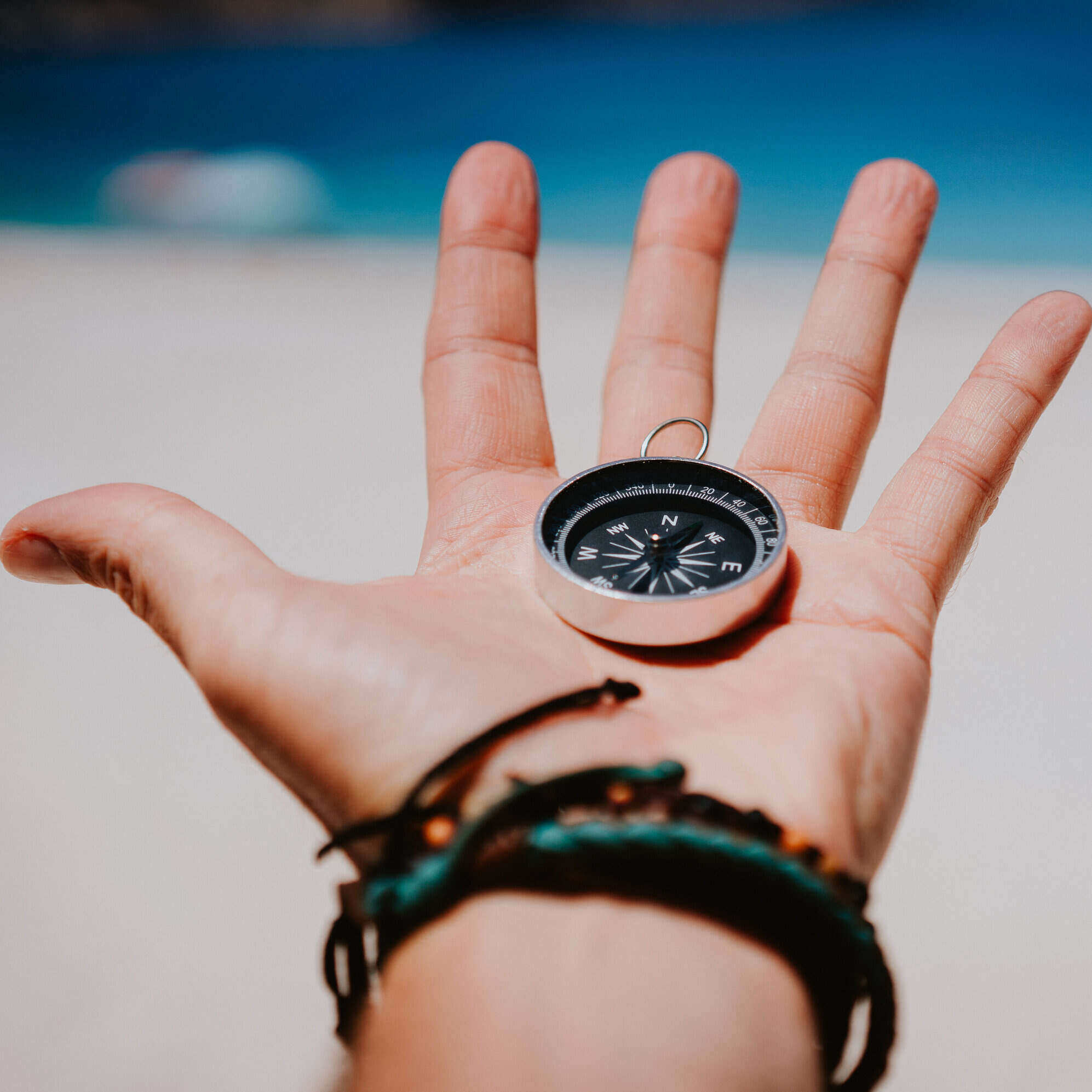 Open palm with stretched fingers holding black metal compass against white sandy beach. Find your way or goal concept. Point of view pov.